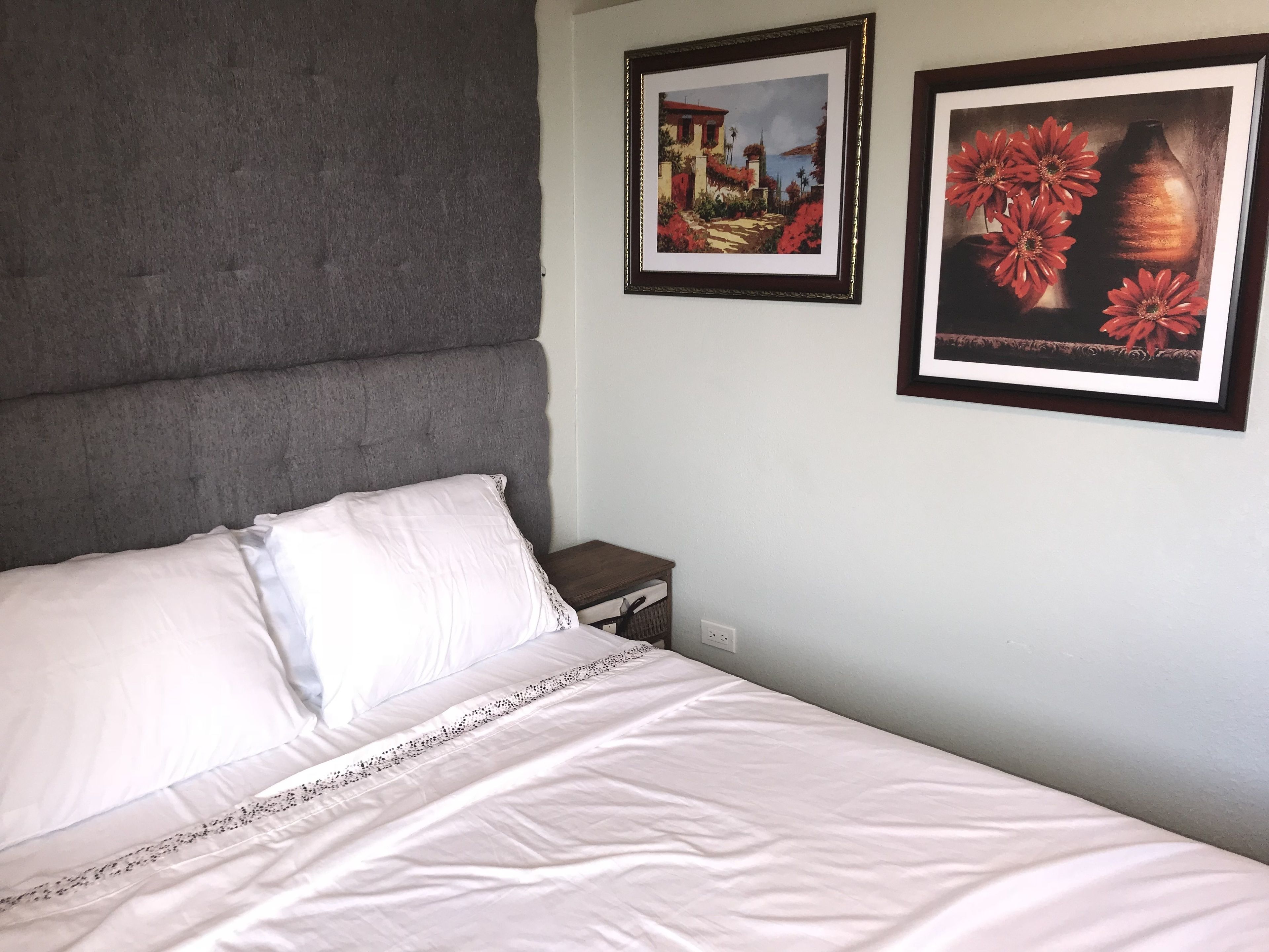 A neatly made bed with white linens and a decorative border, a grey upholstered headboard, and two framed artworks on the wall.