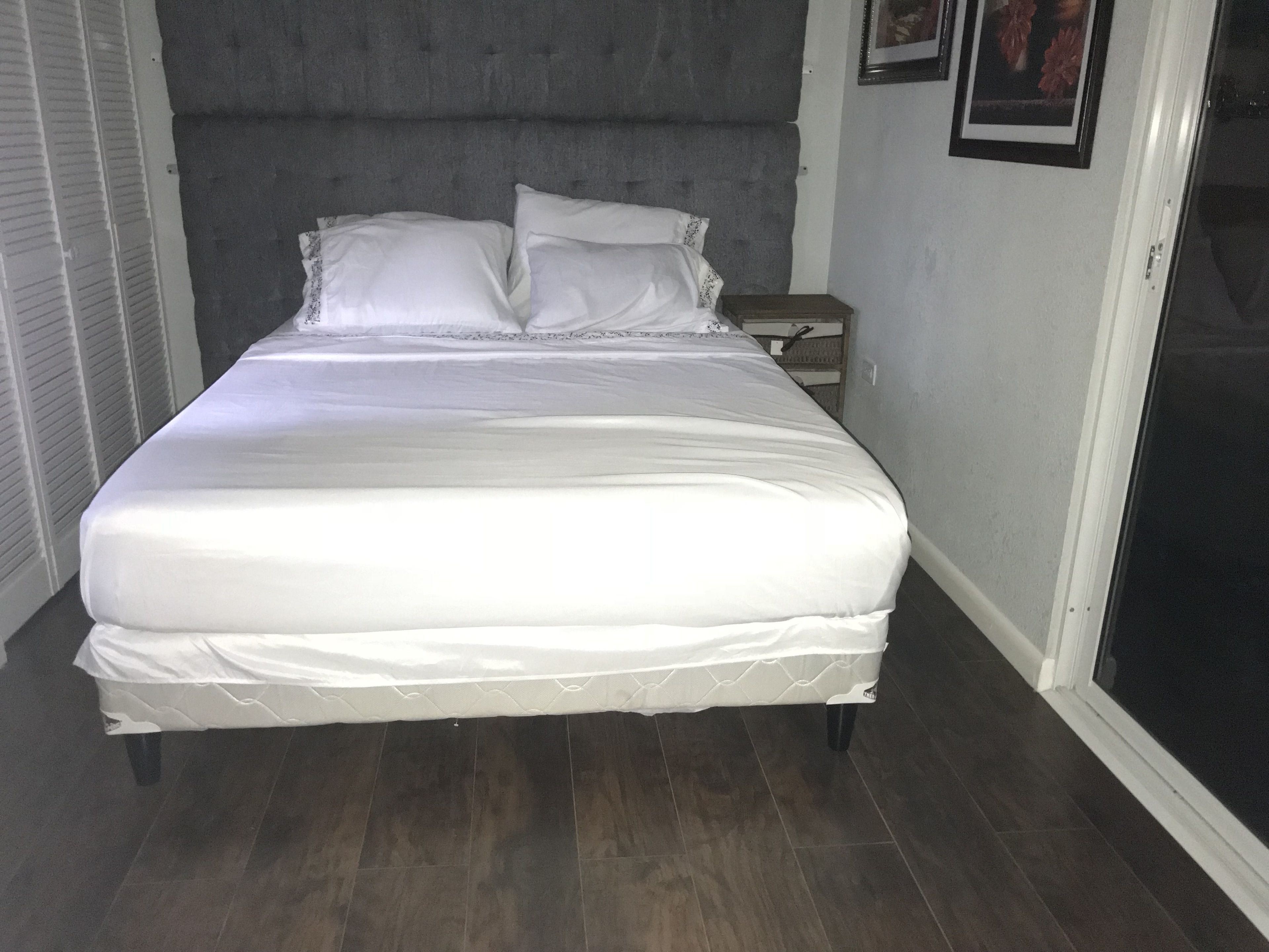 Ein ordentlich bezogenes Bett mit weißer Bettwäsche in einem Zimmer mit dunklem Holzboden und einem grauen, gepolsterten Kopfteil.