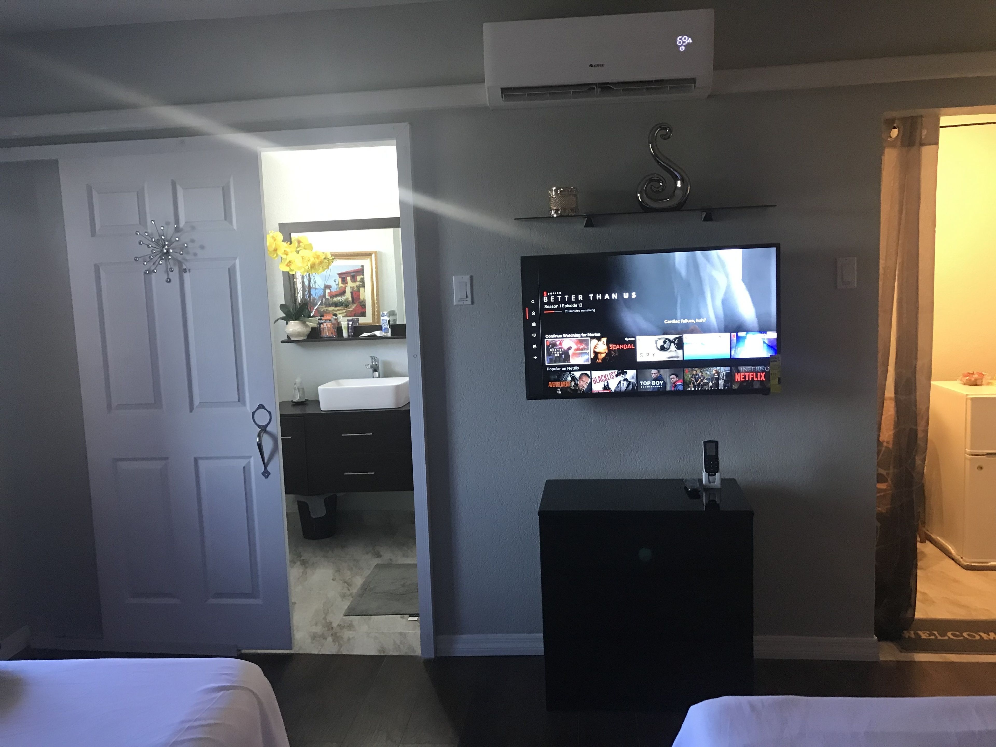 A hotel room with a TV displaying a Netflix interface, a bathroom with a sink and mirror, and a door leading to another room.