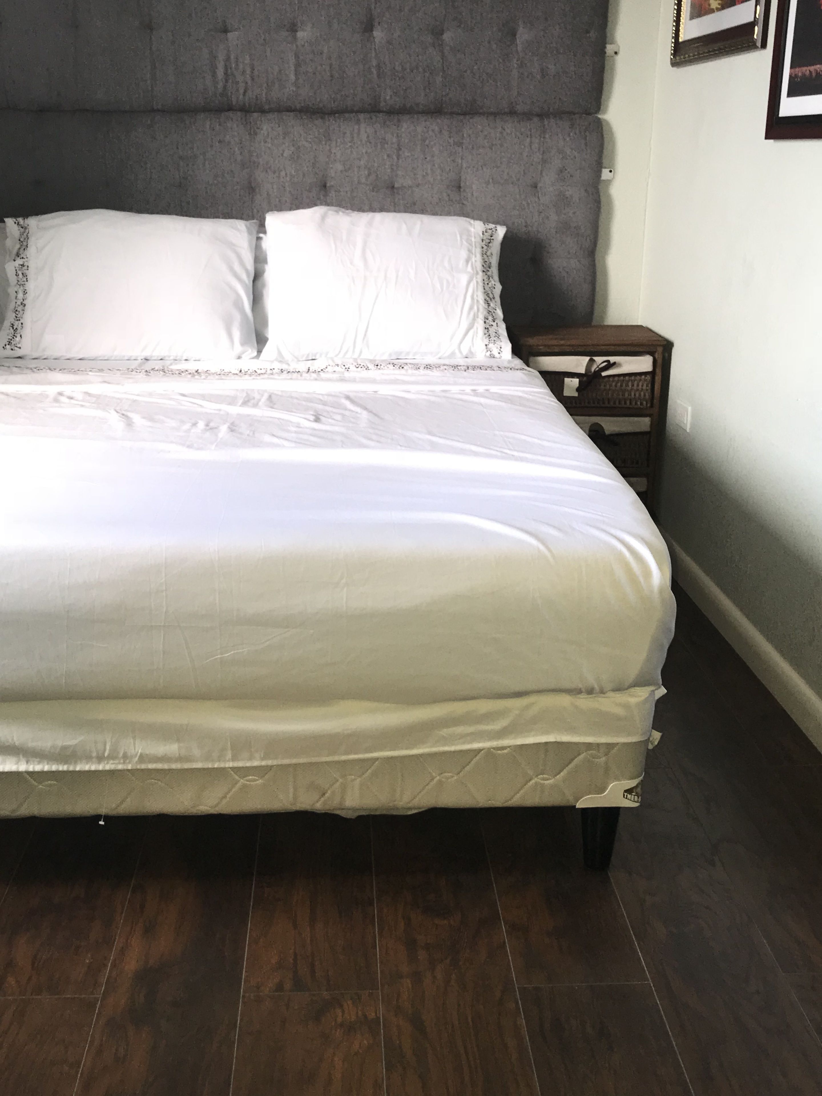 A neatly made bed with white linens and a grey headboard in a room with dark wooden flooring.