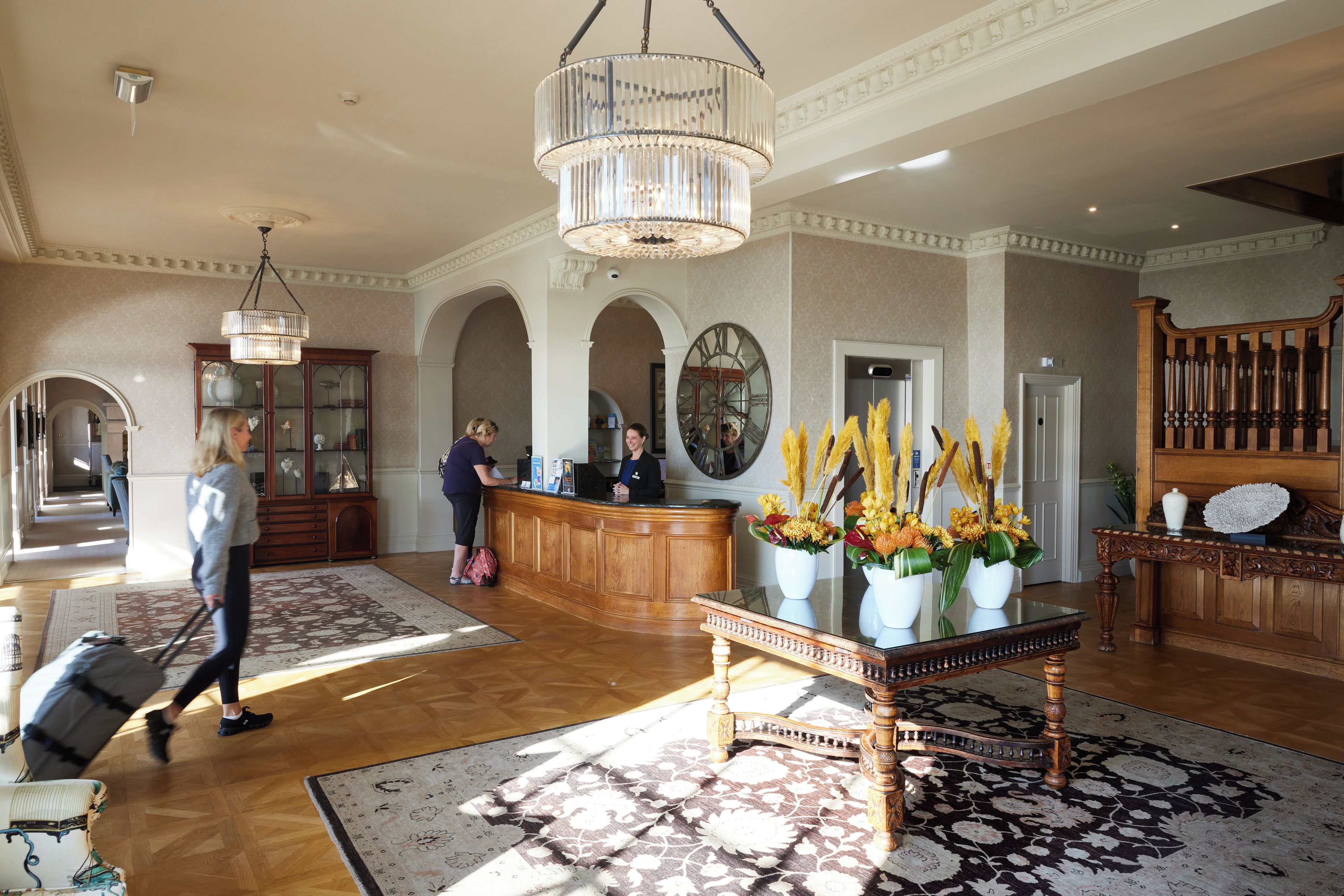 A lobby with a reception desk, a chandelier, a large mirror, and a wooden staircase.