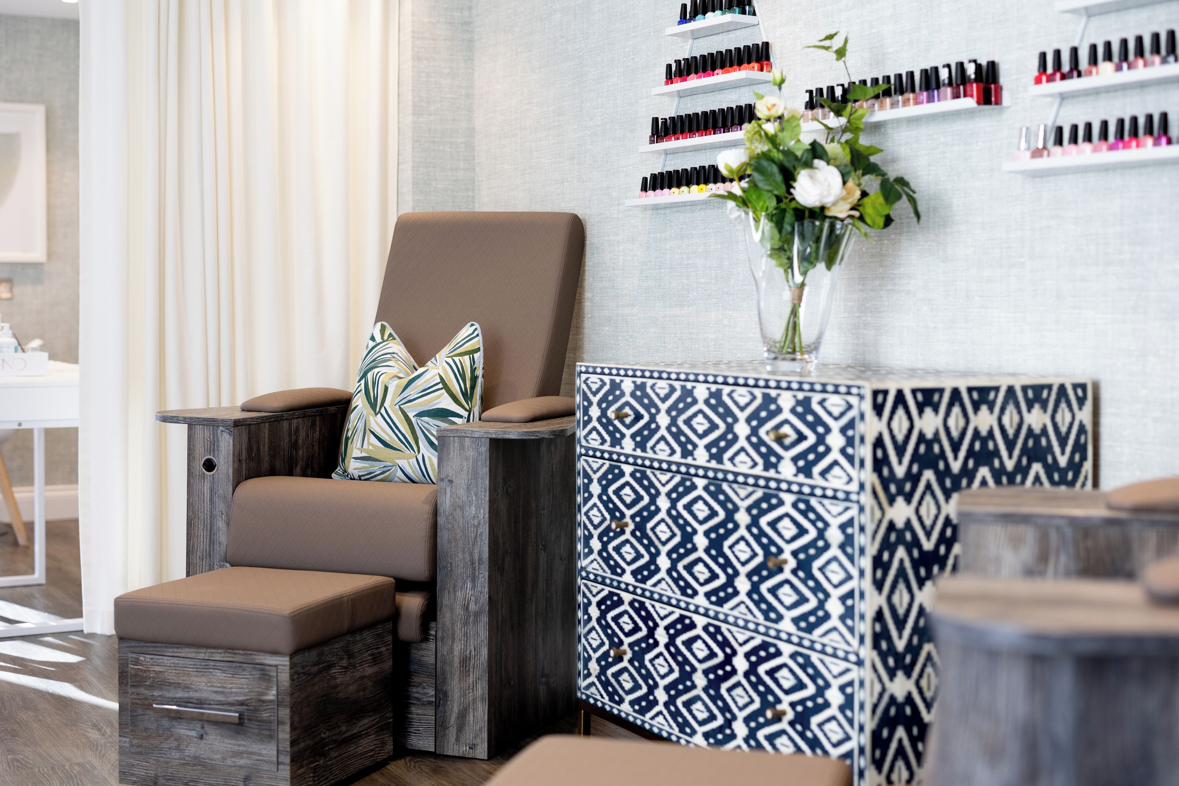 Un sillón de salón con cojín, una mesita auxiliar con un florero estampado y una pared con esmaltes de uñas.
