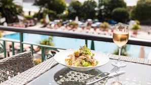Una mesa de comedor con un plato de comida y una copa de vino, con vista a un área de piscina con sillas de descanso y sombrillas.