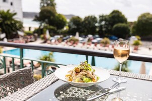 Una mesa de comedor con un plato de comida y una copa de vino, con vista a un área de piscina con sillas de descanso y sombrillas.