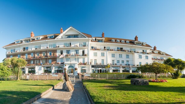 Un grande hotel a più piani con una facciata curva, balconi adornati con fioriere e un ingresso centrale con un viale che conduce alle porte principali.