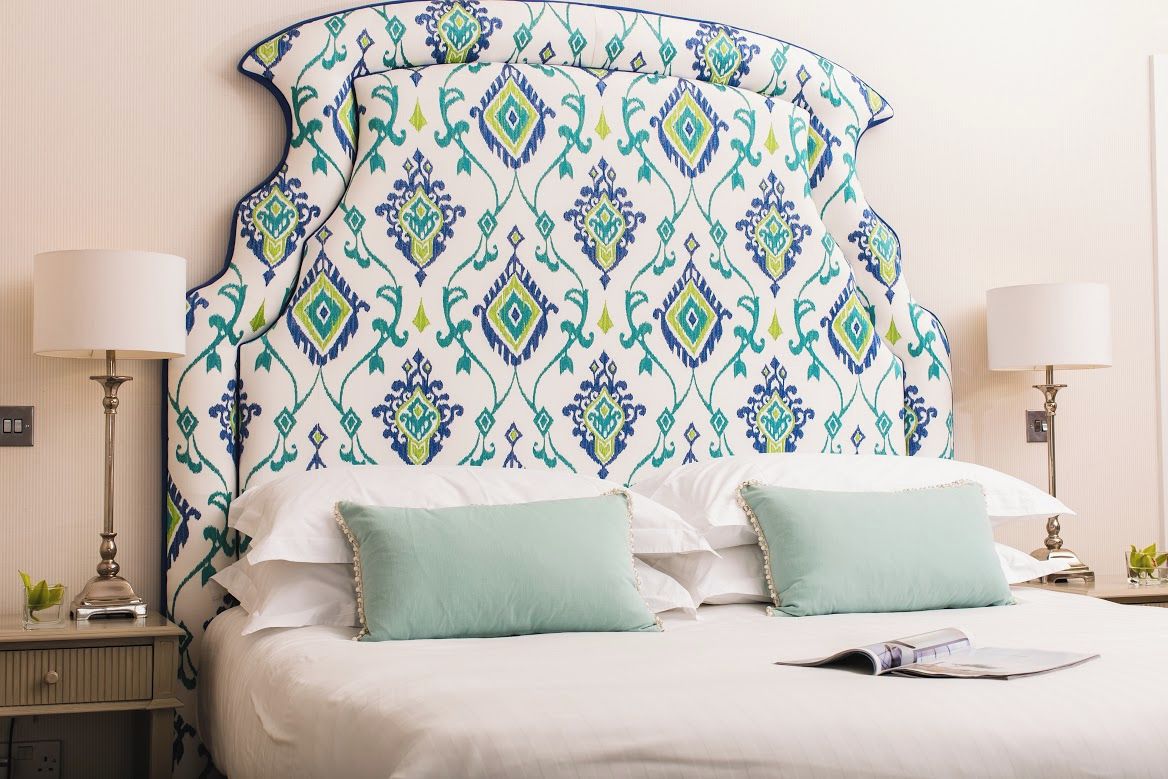 A neatly made bed with a patterned headboard, two bedside lamps, and a bedside table with a lamp and a small plant.