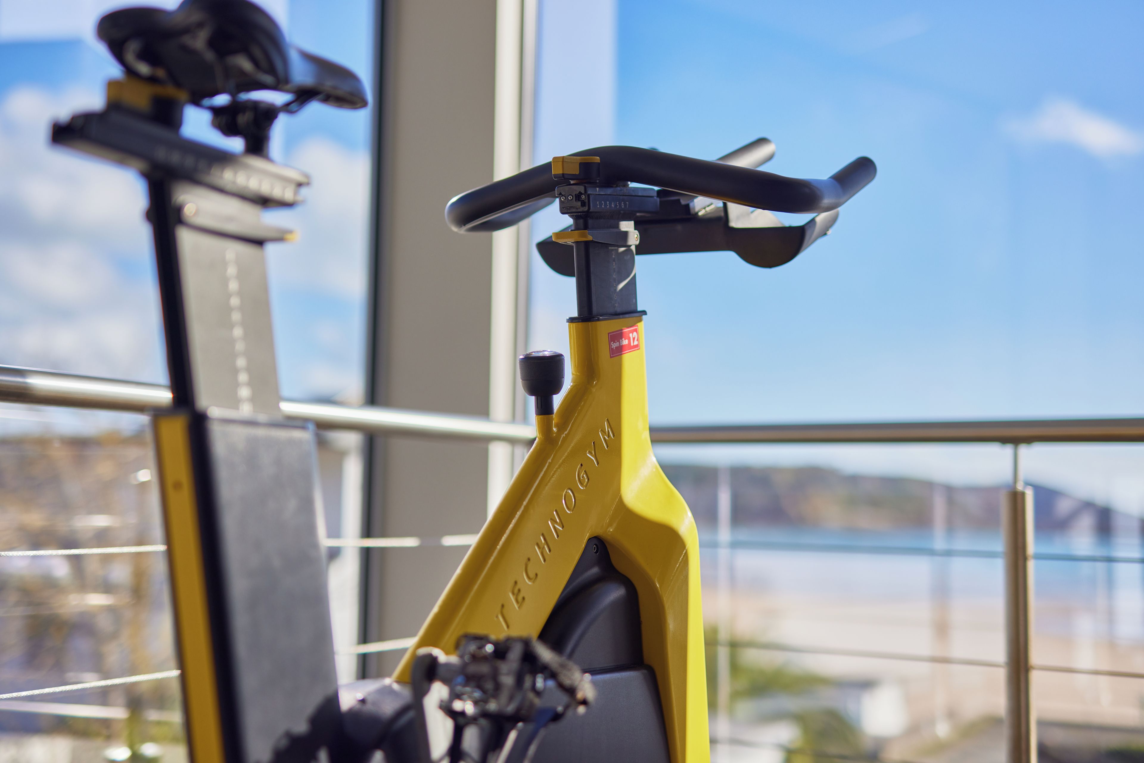 En gul motionscykel med en digital display, placerad inomhus med utsikt över en sjö eller ett annat vattendrag och berg genom ett fönster.