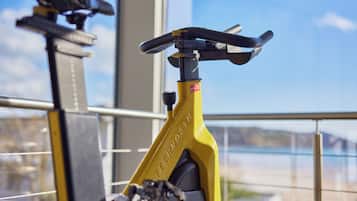 En gul motionscykel med en digital display, placerad inomhus med utsikt över en sjö eller ett annat vattendrag och berg genom ett fönster.