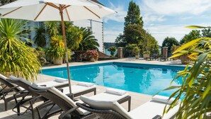 Una piscina con lettini e ombrellone, immersa nel verde e affiancata da un edificio.