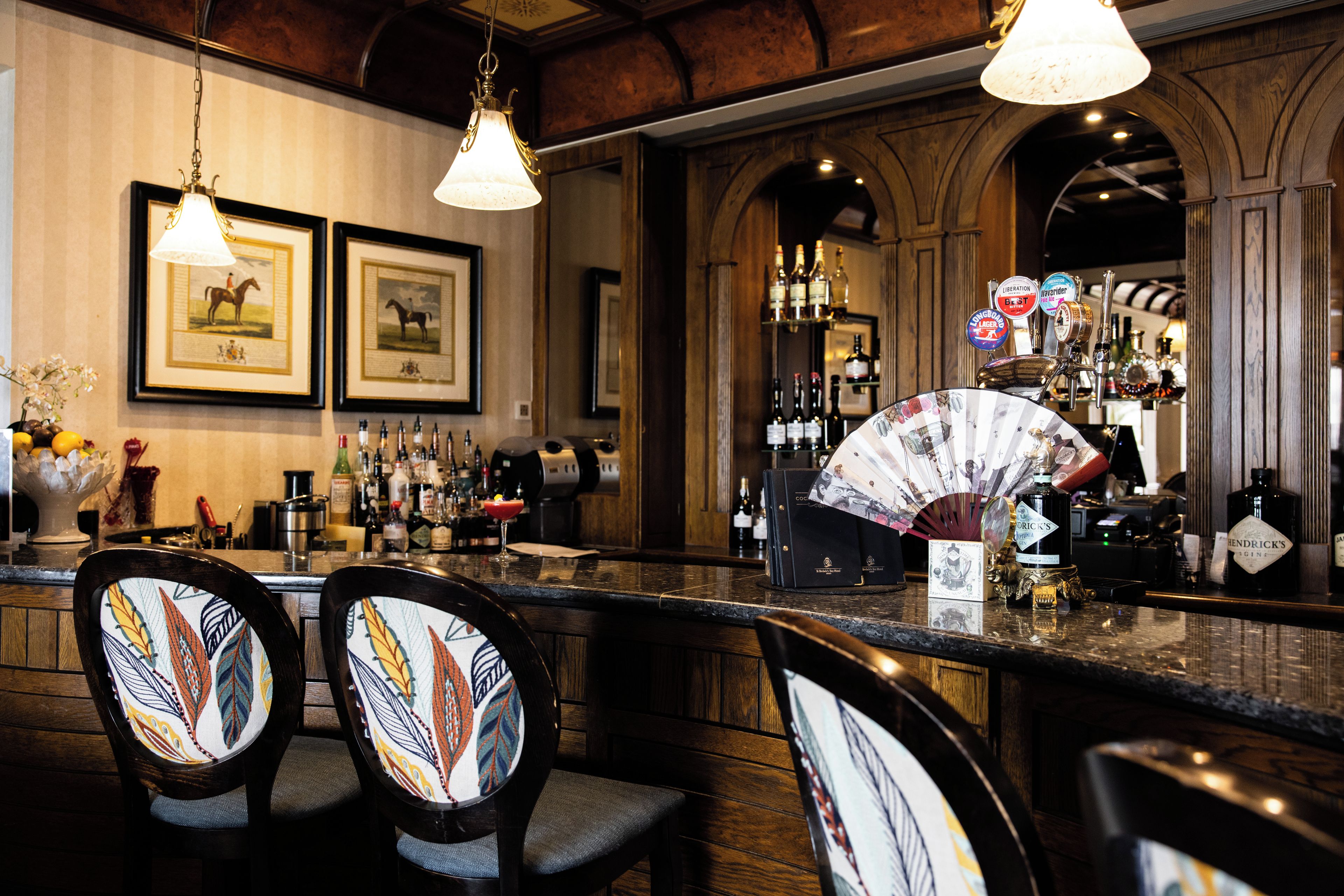 A bar area with wooden chairs, a fan, and a variety of bottles and glasses.
