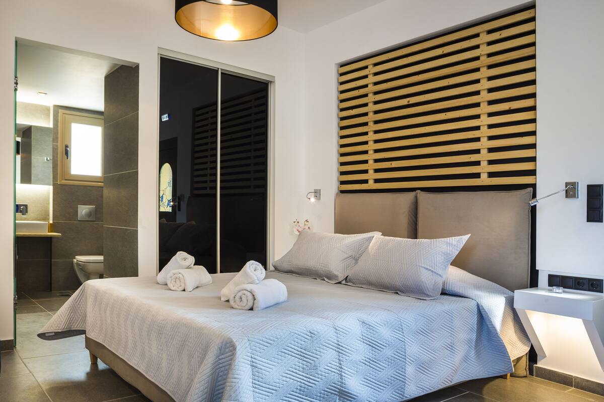 Une chambre d’hôtel moderne, dotée d’un grand lit, d’une table de chevet, d’un miroir et d’une salle de bain visible à travers une porte ouverte.