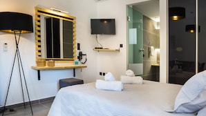 Une chambre d’hôtel moderne avec un grand lit, un téléviseur fixé au mur, une table de chevet avec une lampe et une salle de bain visible à travers une porte en verre.