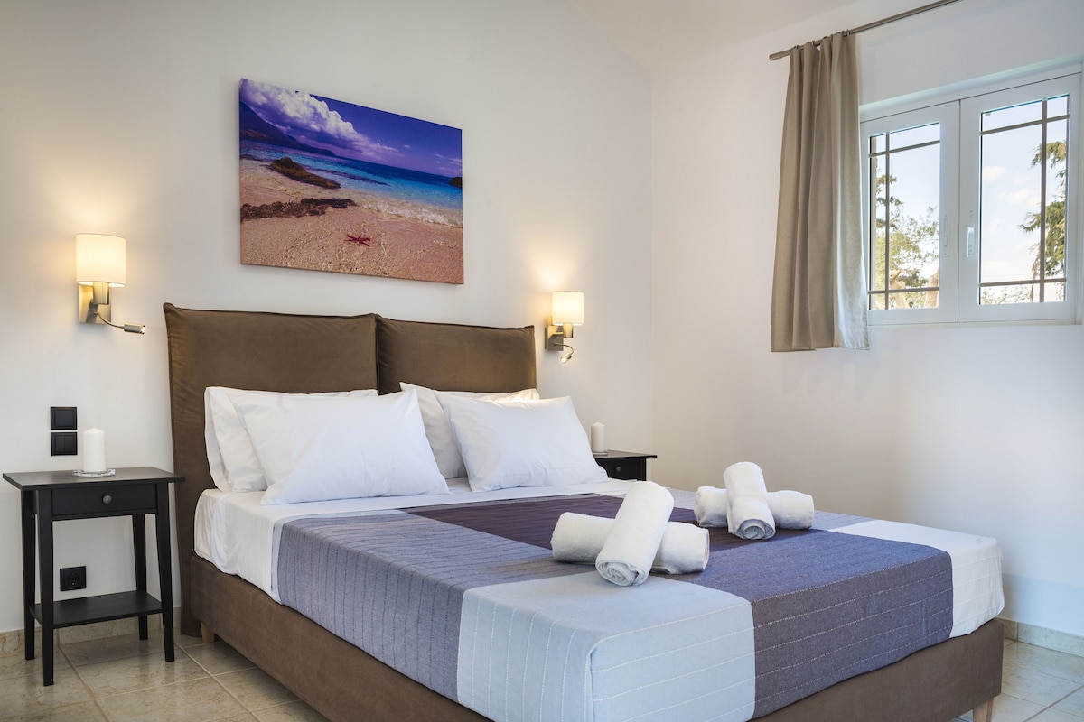 Un lit bien fait, avec une literie blanche et bleue, deux tables de chevet avec des lampes, un tableau sur le thème de la plage accroché au mur et une fenêtre avec des rideaux.