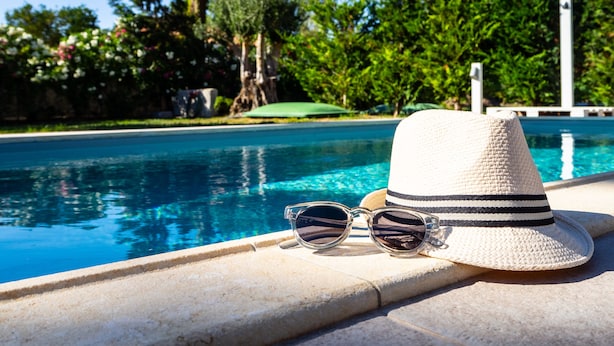 Une scène au bord de la piscine, avec un chapeau et des lunettes de soleil posés sur un rebord en pierre.