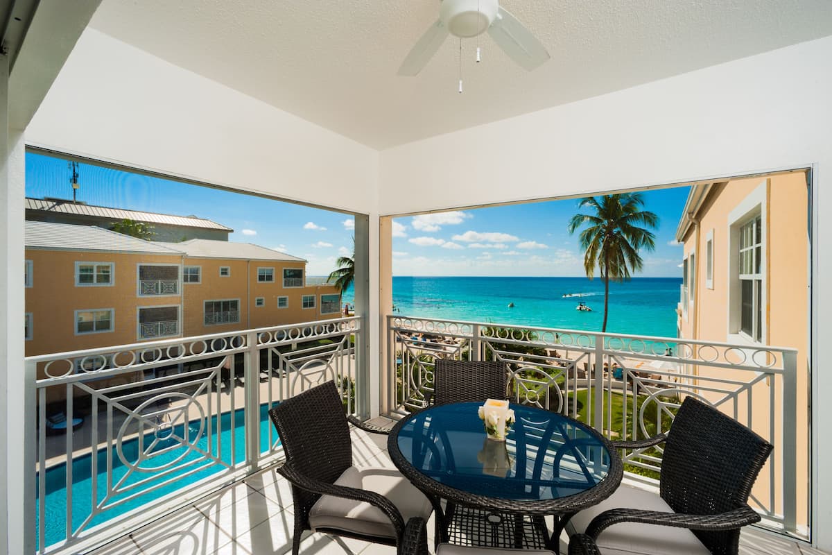 A balcony with a glass table and chairs, overlooking a swimming pool and a beach with palm trees.