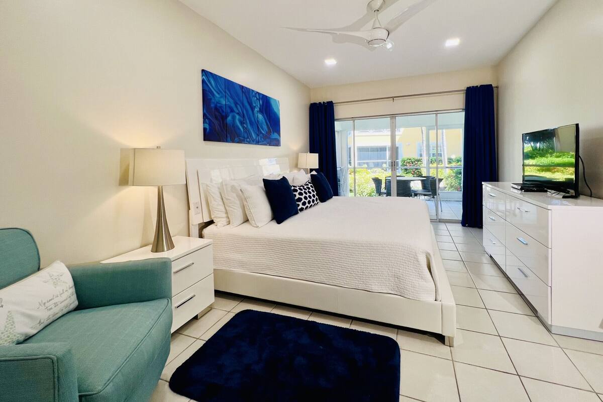 A modern bedroom with a large bed, a flat-screen TV, a ceiling fan, and a view of a balcony with outdoor furniture.
