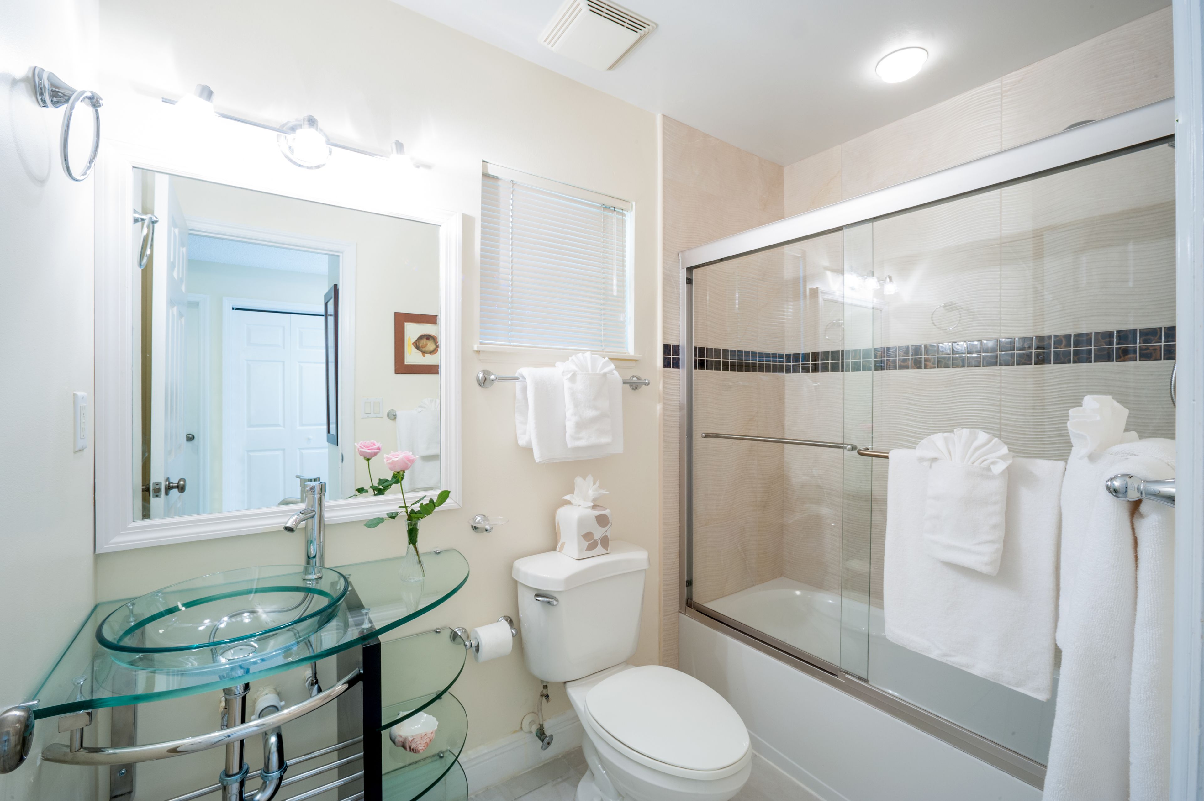 Un baño moderno con un lavabo de vidrio, un inodoro, una ducha con cerramiento de vidrio y un espejo con luces arriba.