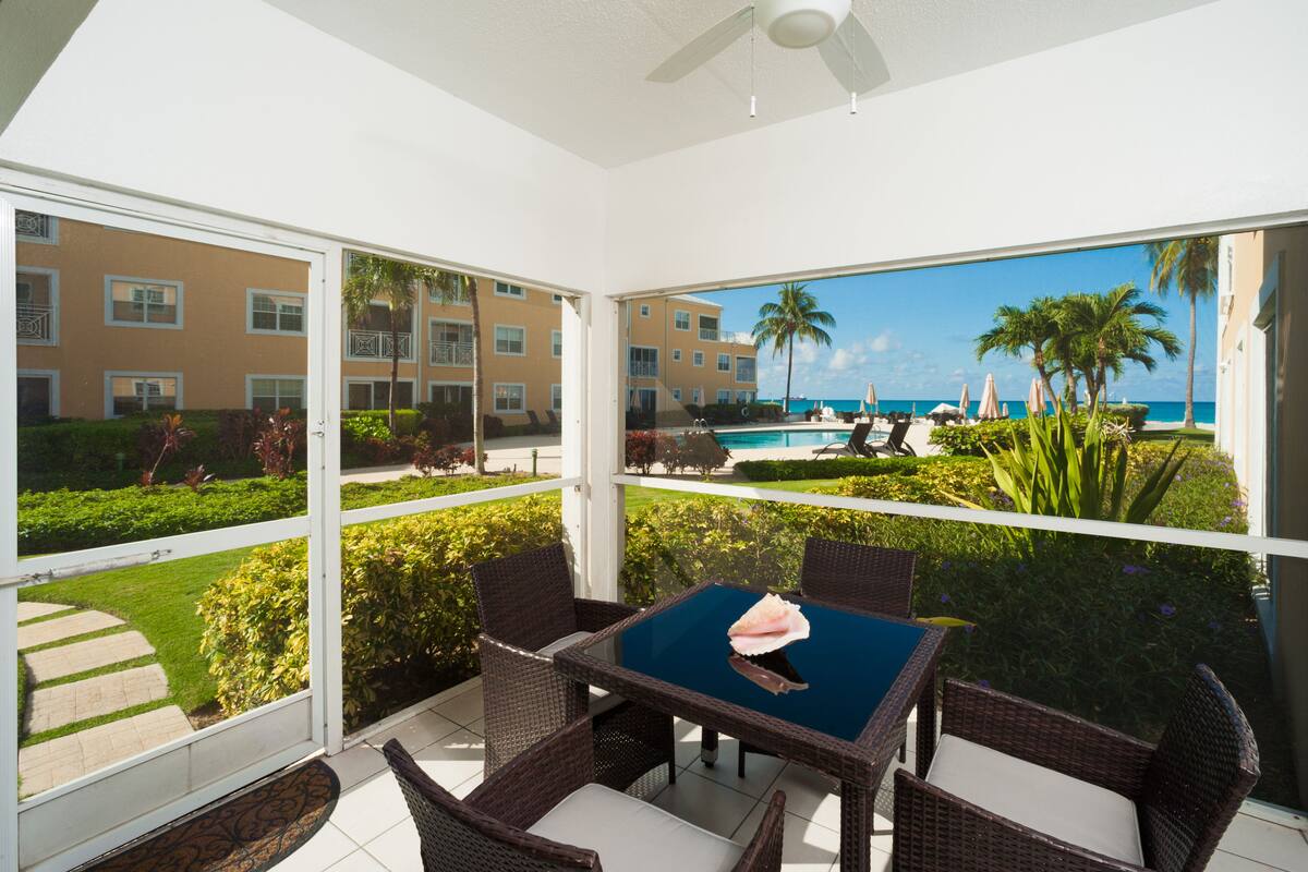 A balcony with wicker furniture, a glass table, and a view of a pool and palm trees.