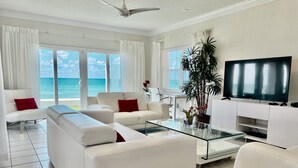 A modern living room with a large TV, white sofas, a glass coffee table, and a view of the ocean. - Regal Beach Club (Seven Mile Beach)