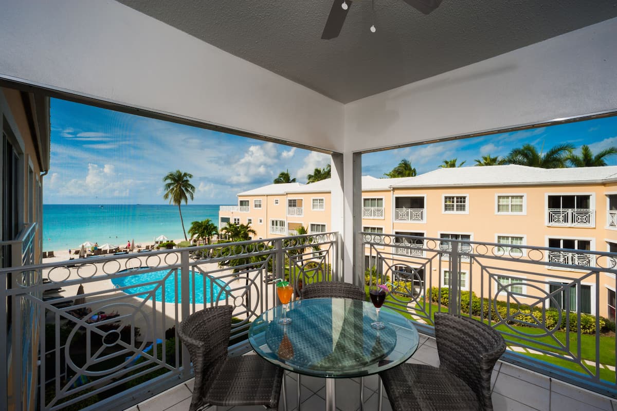 A balcony with a glass table and wicker chairs, overlooking a pool and beach.