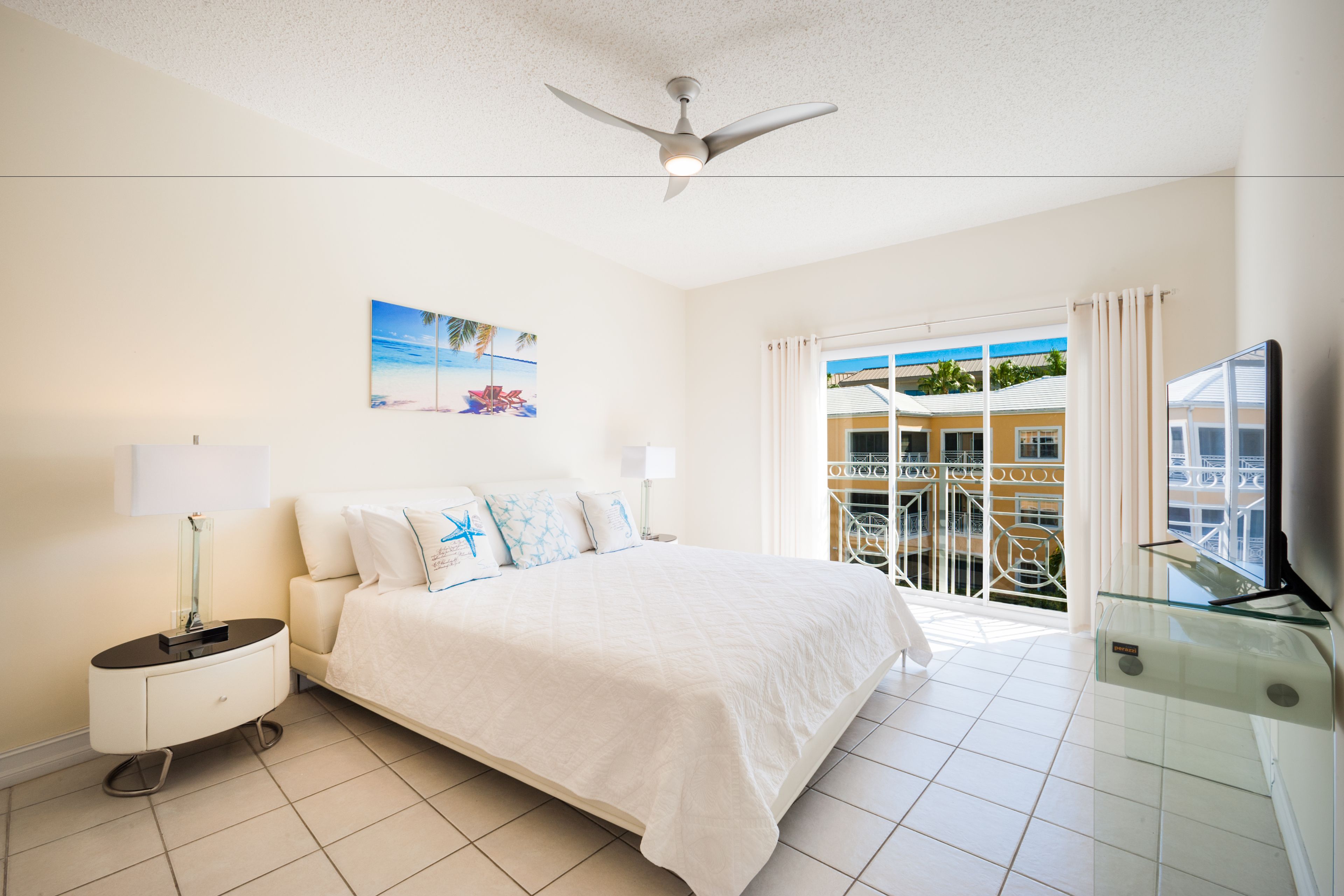 A hotel room with a bed, a nightstand, a TV, a ceiling fan, and a balcony with a view of buildings and palm trees.