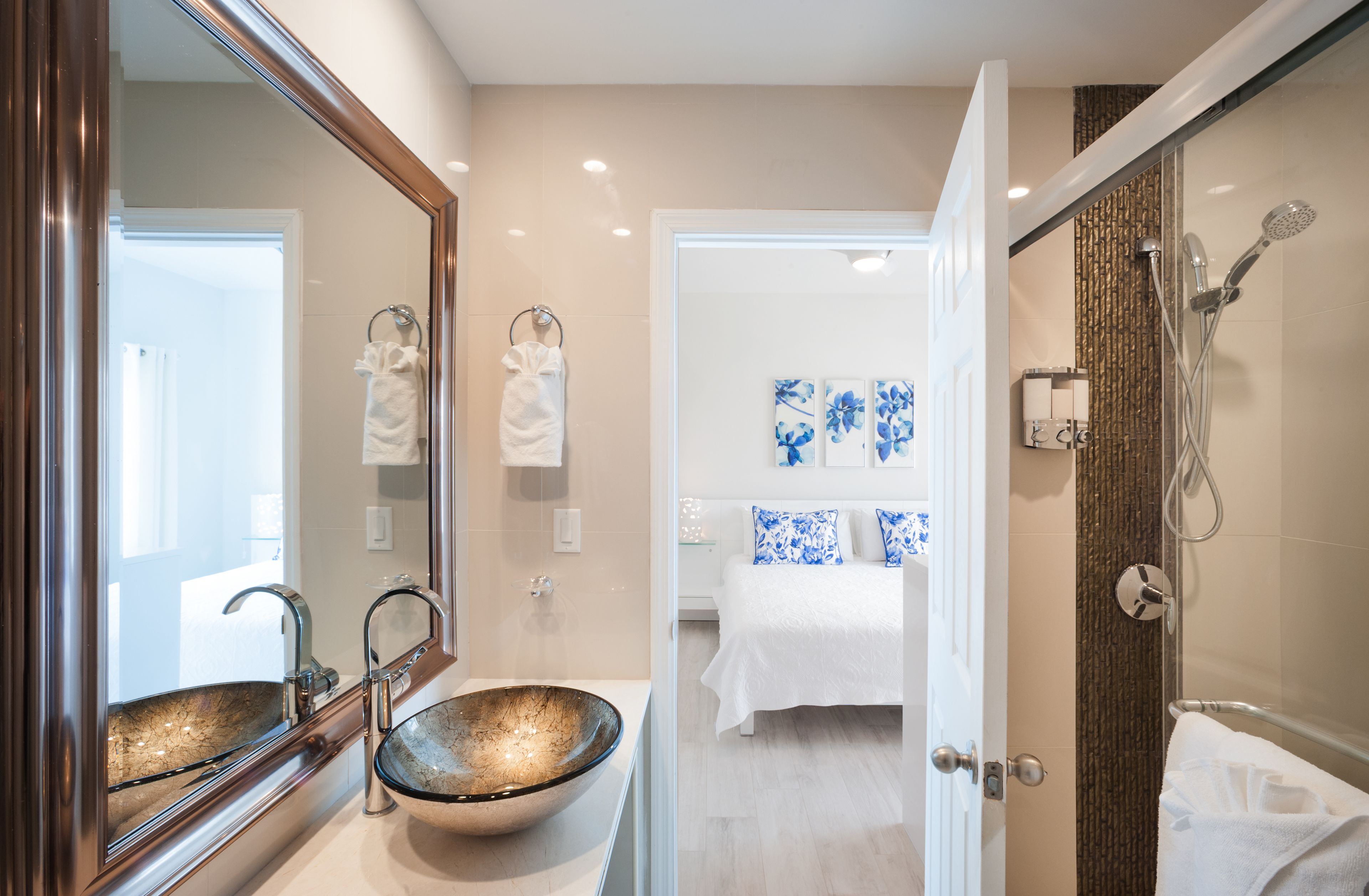 A modern bathroom with a large mirror, a glass bowl sink, a shower with a handheld showerhead, and a bed with blue floral pillows in the adjacent room.