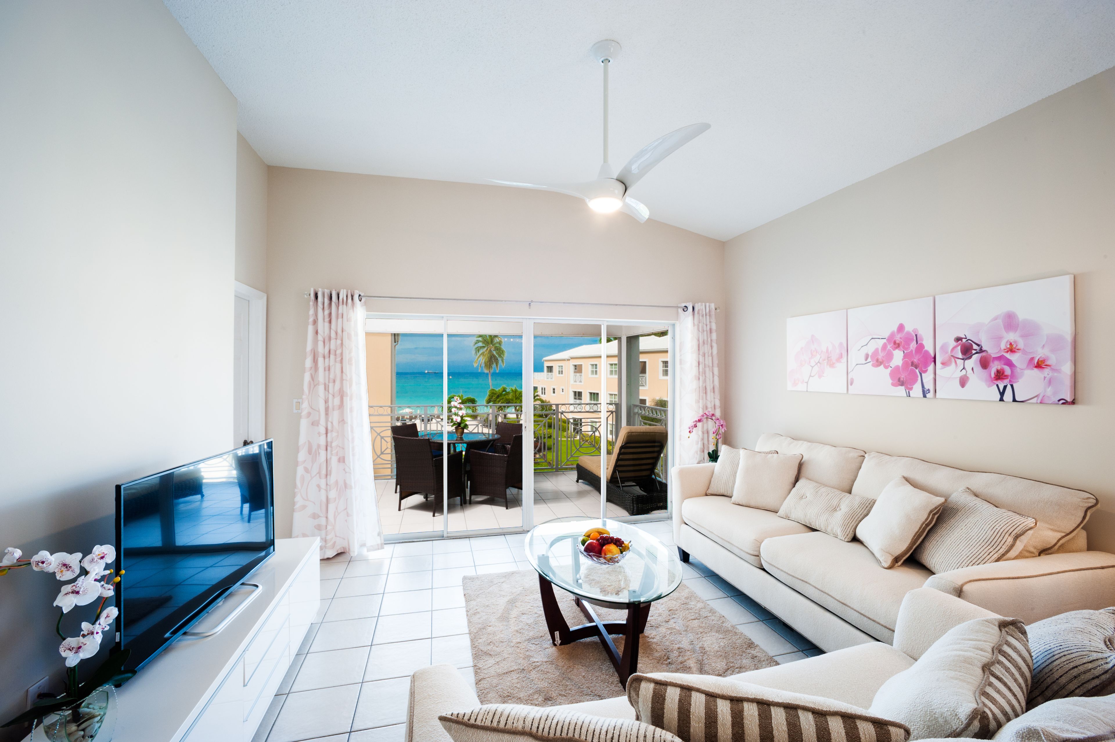 A modern living room with a flat-screen TV, a glass coffee table, a beige sofa, and a balcony with a view of the beach and palm trees.