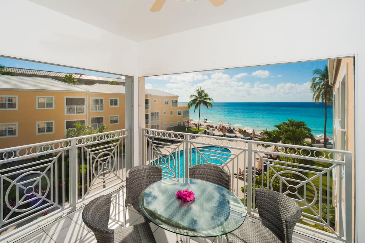 A balcony with wicker chairs and a glass table, overlooking a pool and beach with palm trees and a building in the background.