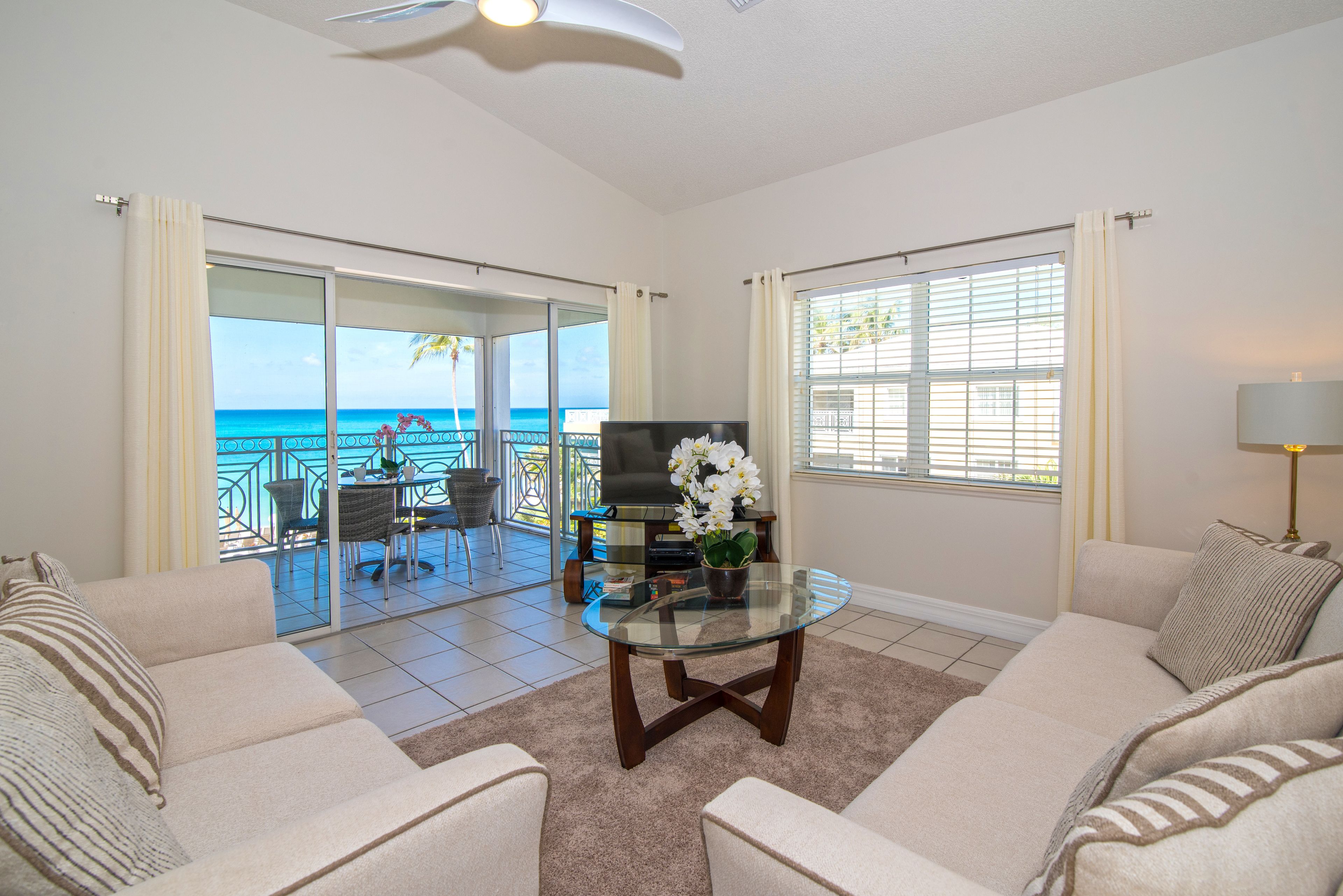 A living room with a glass coffee table, two sofas, a TV, and a view of the ocean through large sliding doors.