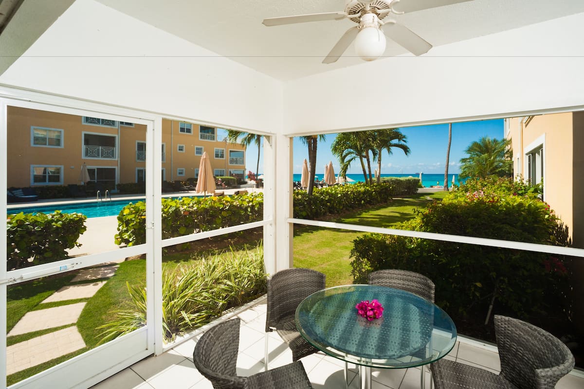 A glass-enclosed balcony with a pool view, wicker chairs, and a glass table with a flower centerpiece.
