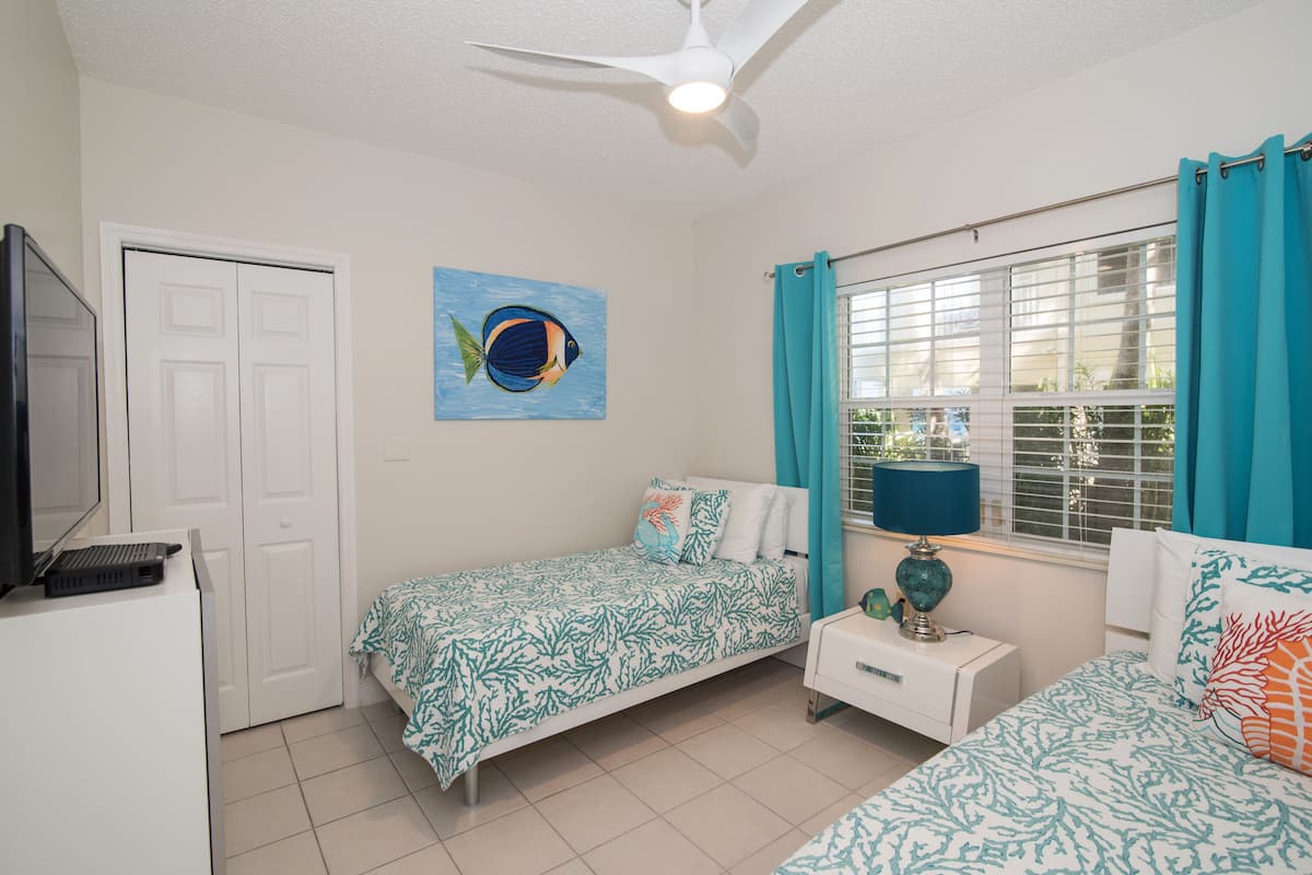 A bedroom with two beds, a ceiling fan, a window with curtains, a nightstand, and a wall painting.