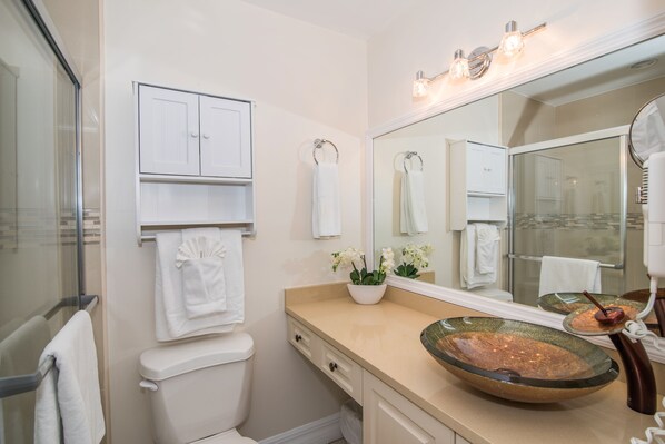 A bathroom with a glass shower enclosure, a beige vanity with a large mirror, a white toilet, and a decorative bowl sink.