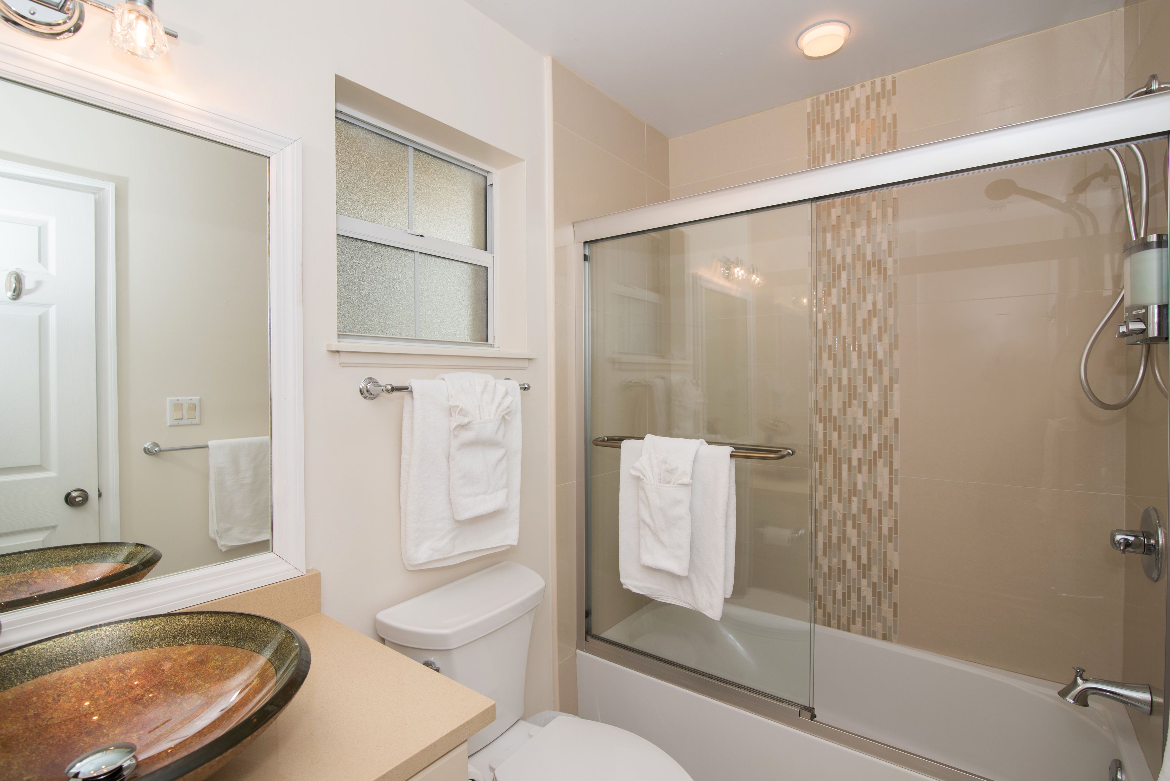 Une salle de bain avec une douche à parois de verre, des toilettes blanches et un grand miroir au-dessus d’un lavabo agrémenté d’un pare-feu en mosaïque.