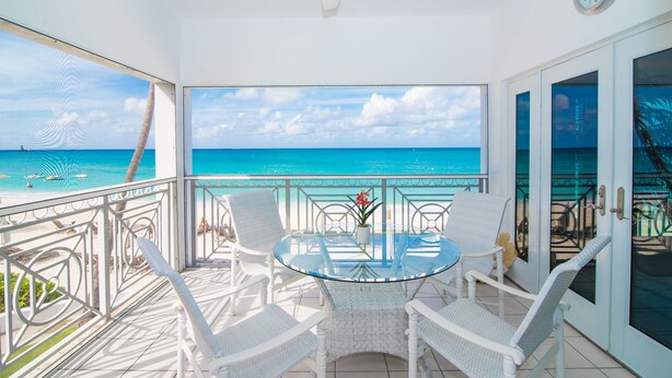 A balcony with a glass table and white wicker chairs, overlooking a beach with clear blue water and a boat. - Regal Beach Club (Seven Mile Beach)