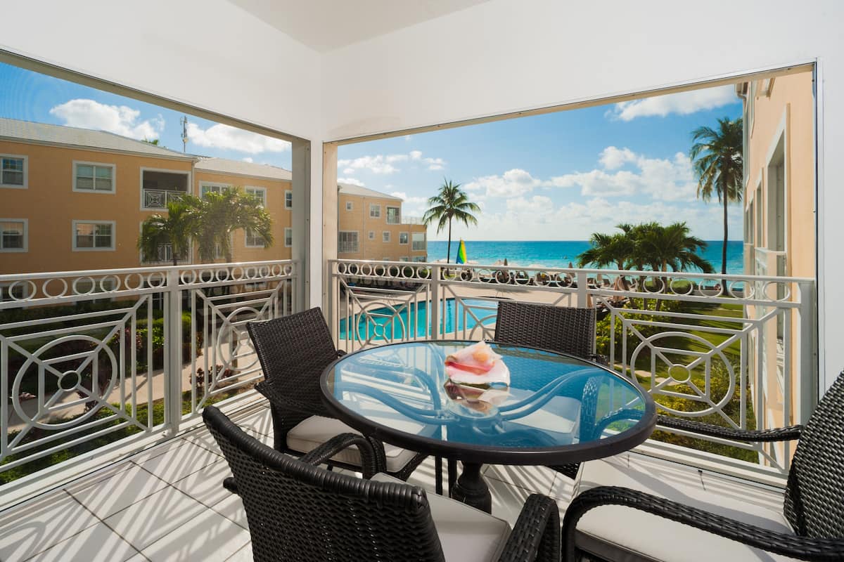 A balcony with wicker furniture, a glass table, and a view of the ocean and buildings.
