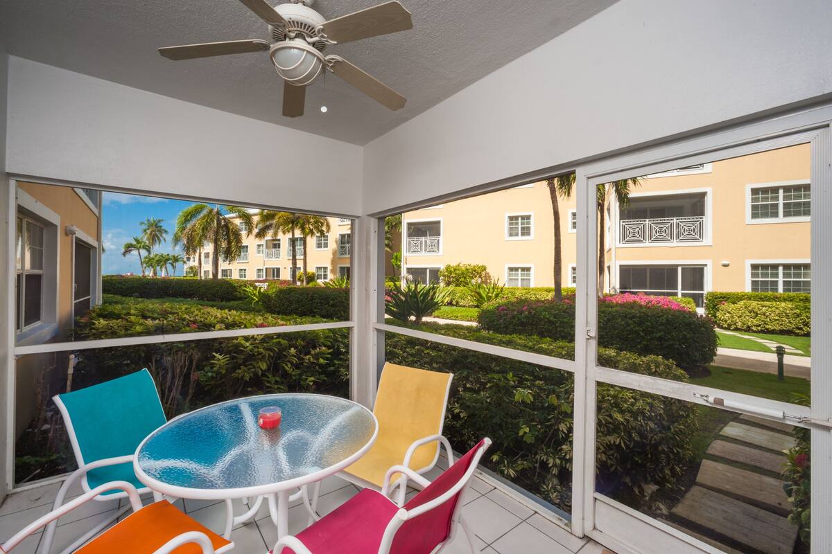 A sunlit room with a glass door leading to a balcony, featuring a table with chairs and a ceiling fan.