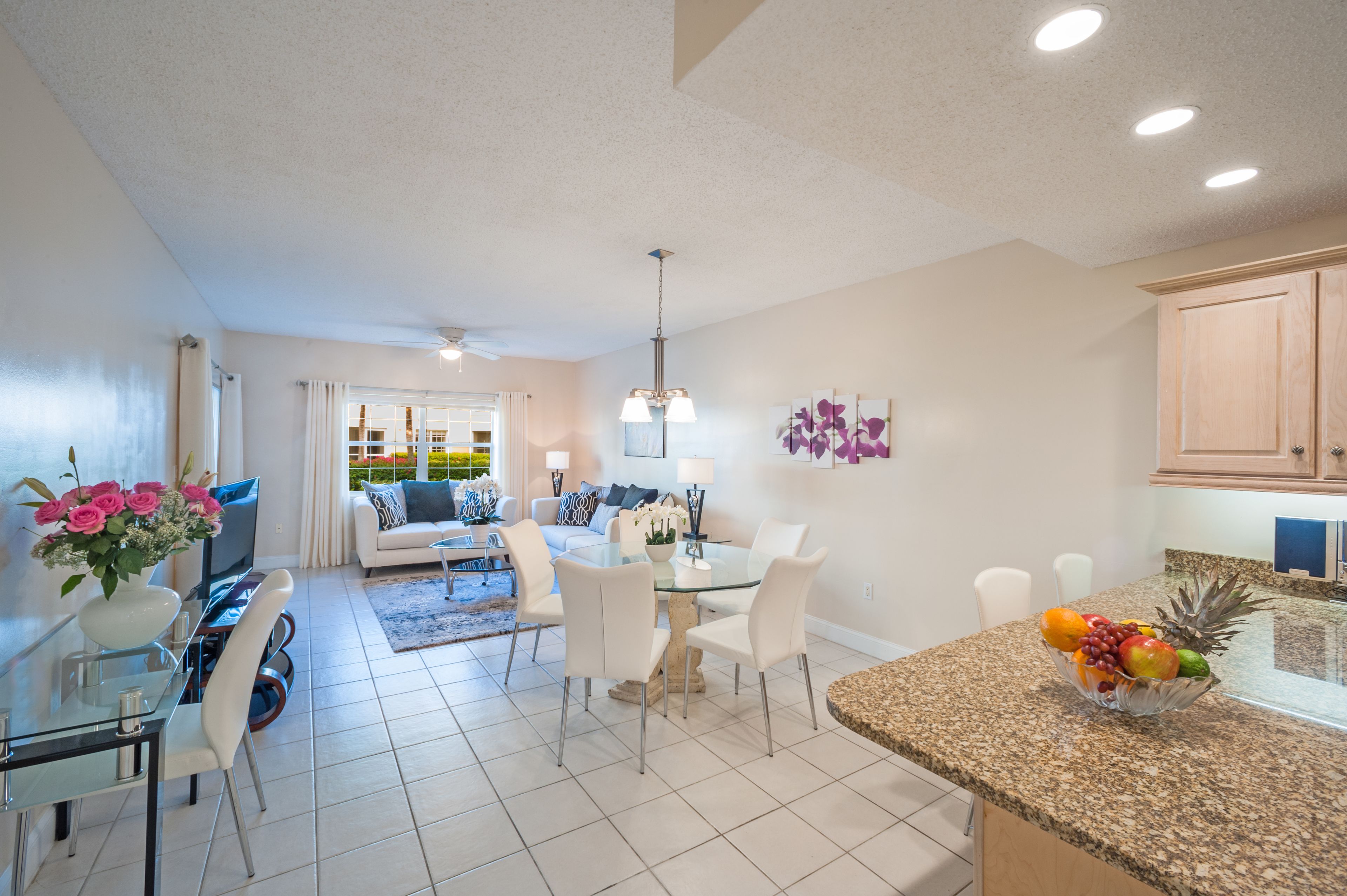 A modern living room with a glass dining table, white sofas, and a kitchen area with granite countertops and wooden cabinets.