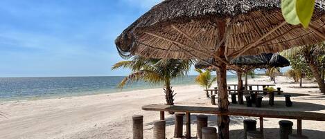 A beach with a thatched umbrella and wooden tables.