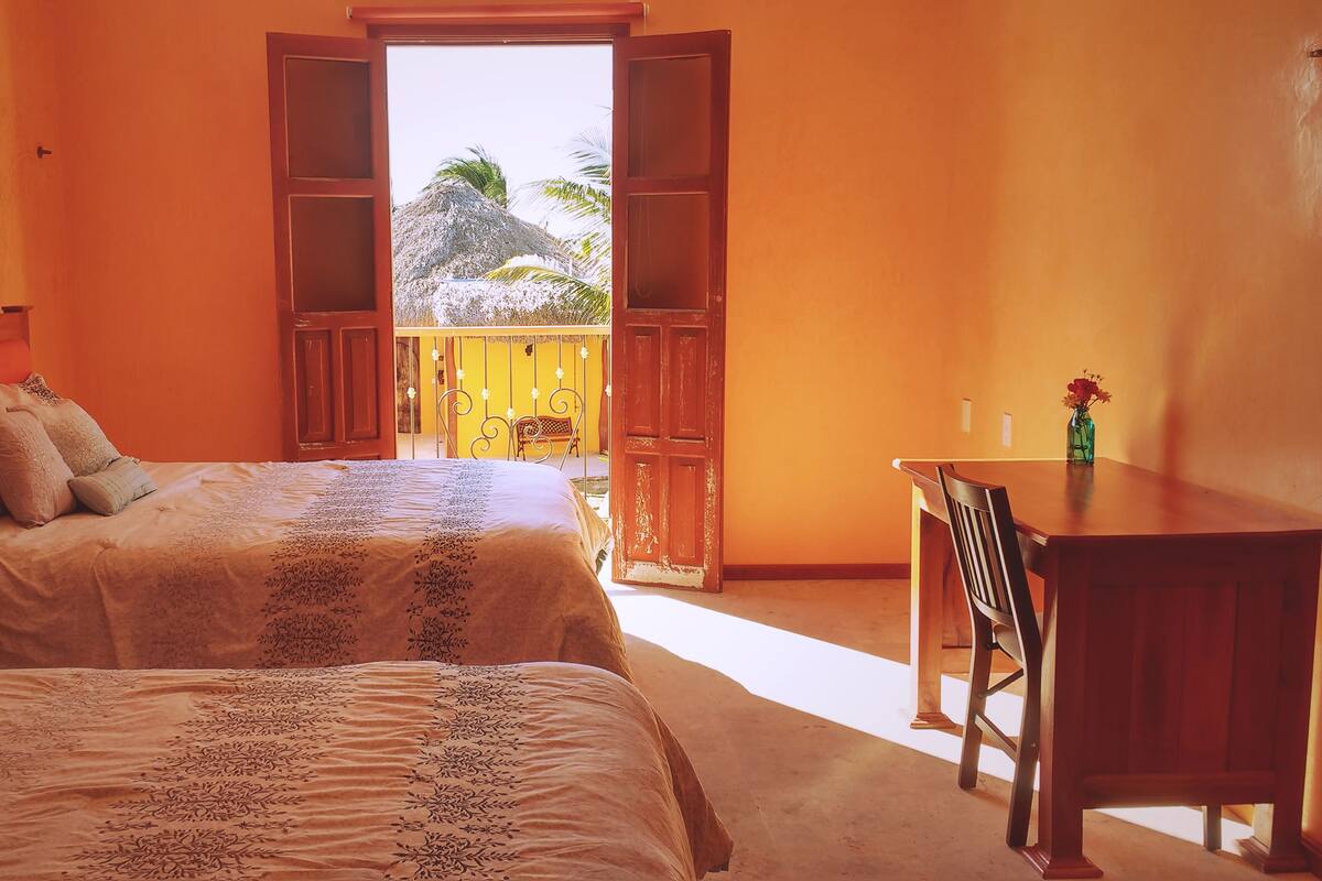 A bedroom with two beds, a wooden desk, and a balcony with a view of palm trees.