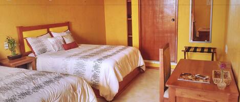 Un dormitorio con dos camas, una mesita de noche de madera con una planta, una silla de madera, una pequeña mesa con un jarrón y un espejo en la pared.