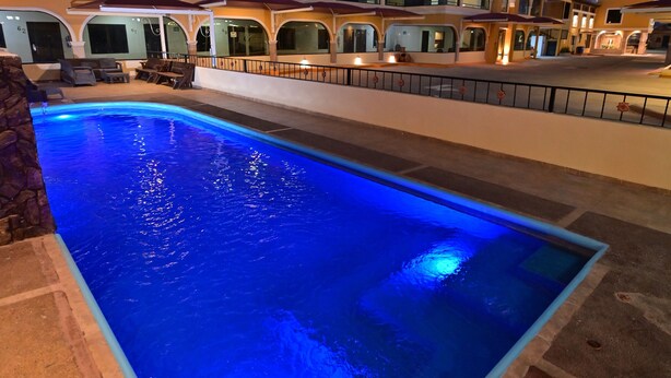 Una piscina iluminada con luz azul, rodeada por un muro de piedra, y un edificio con varios balcones al fondo.