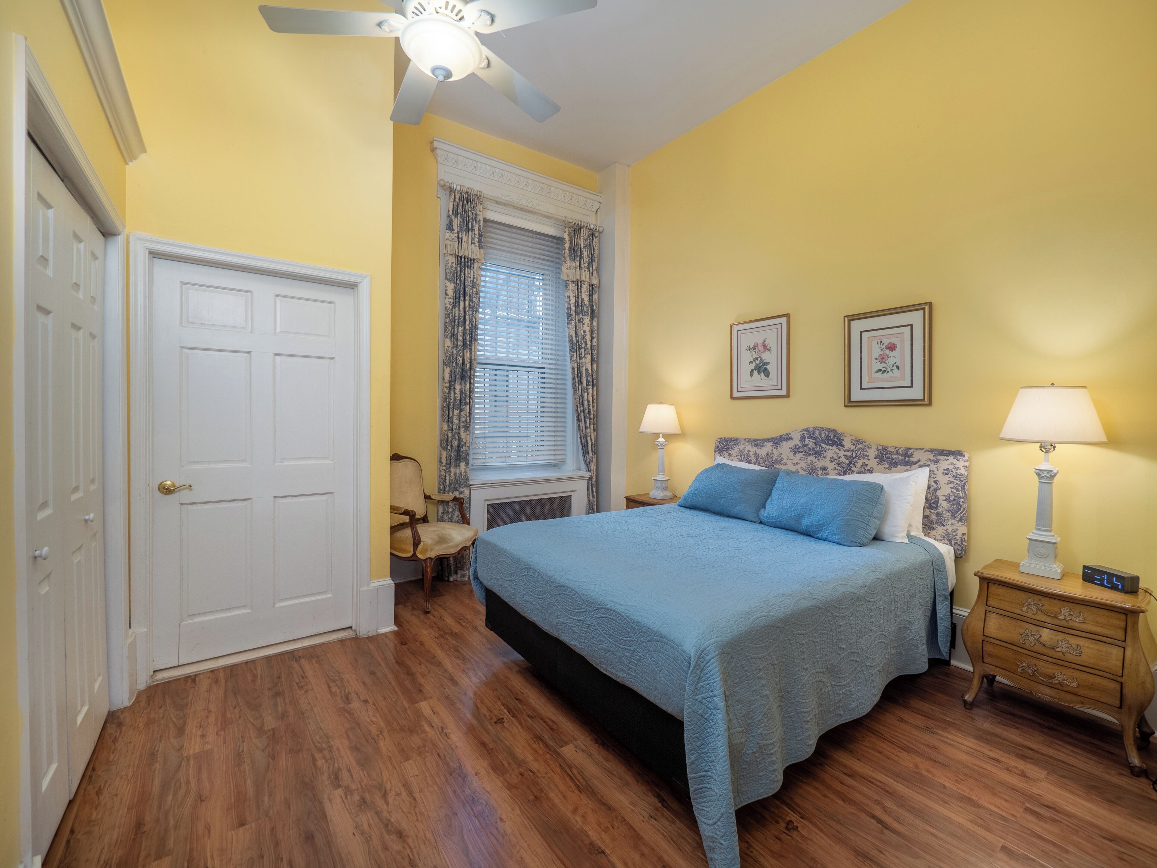 A bedroom with a bed, two bedside tables, a window with curtains, a ceiling fan, and a door.