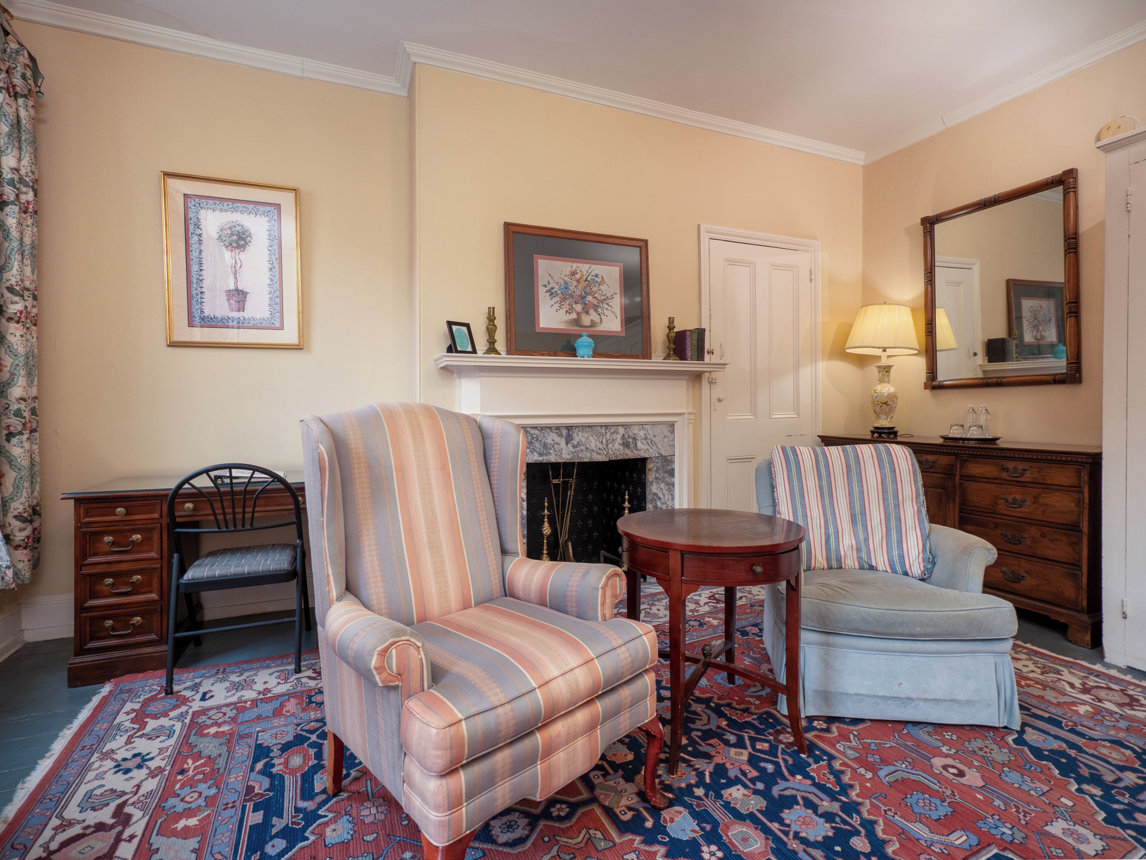 A room with a striped armchair, a blue armchair, a wooden side table, a fireplace, and a large mirror.