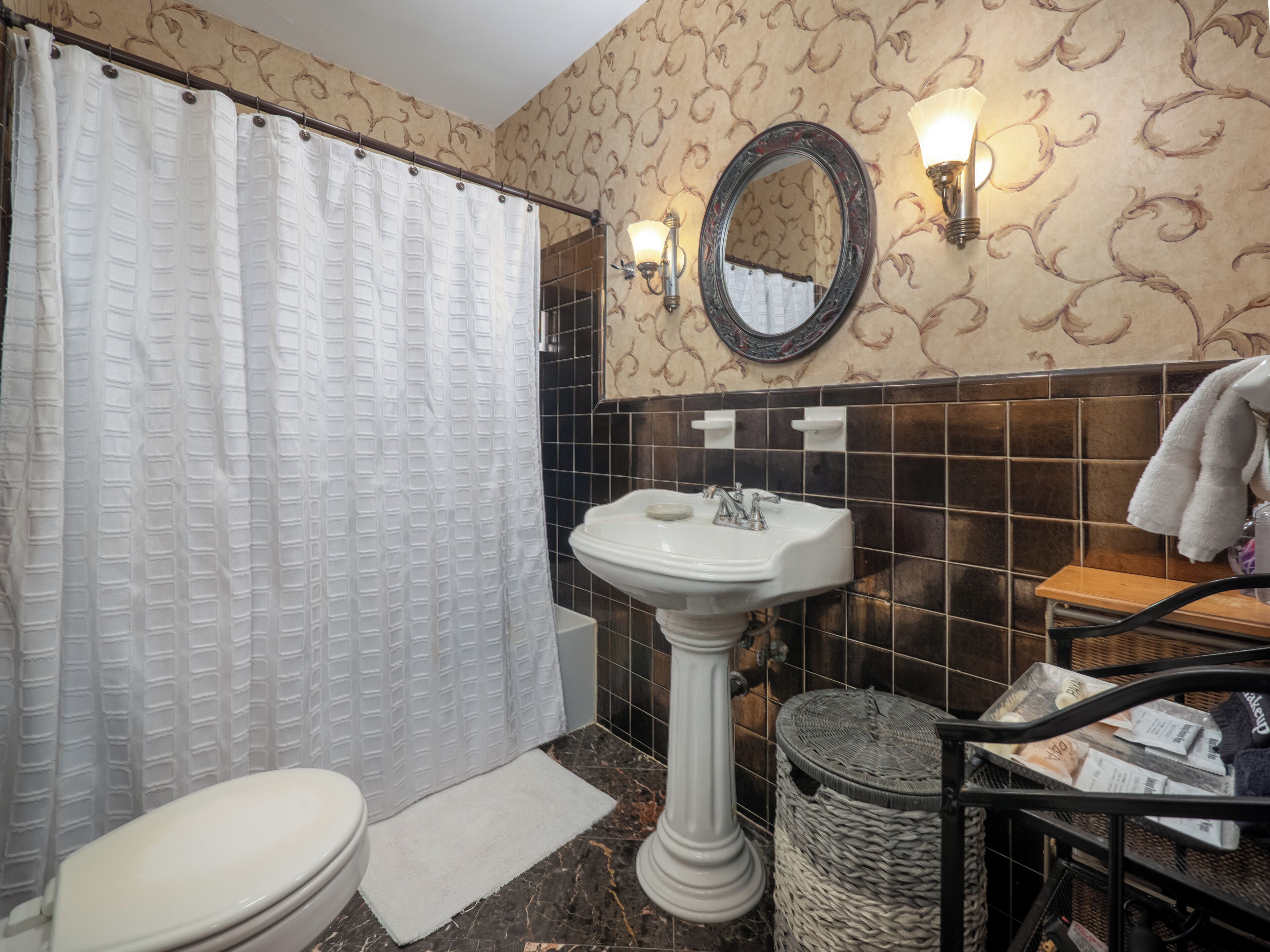 A bathroom with a white toilet, a pedestal sink, a shower curtain, a mirror, and a trash bin.