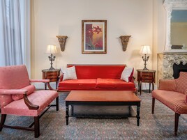 Quarto com sofá vermelho, poltrona combinando, mesa de centro de madeira, duas luminárias fixadas na parede e um quadro emoldurado.
