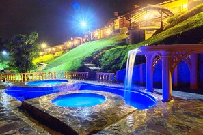 Vista nocturna d'un hotel amb zona de piscina, cascada i escala que puja una collada.
