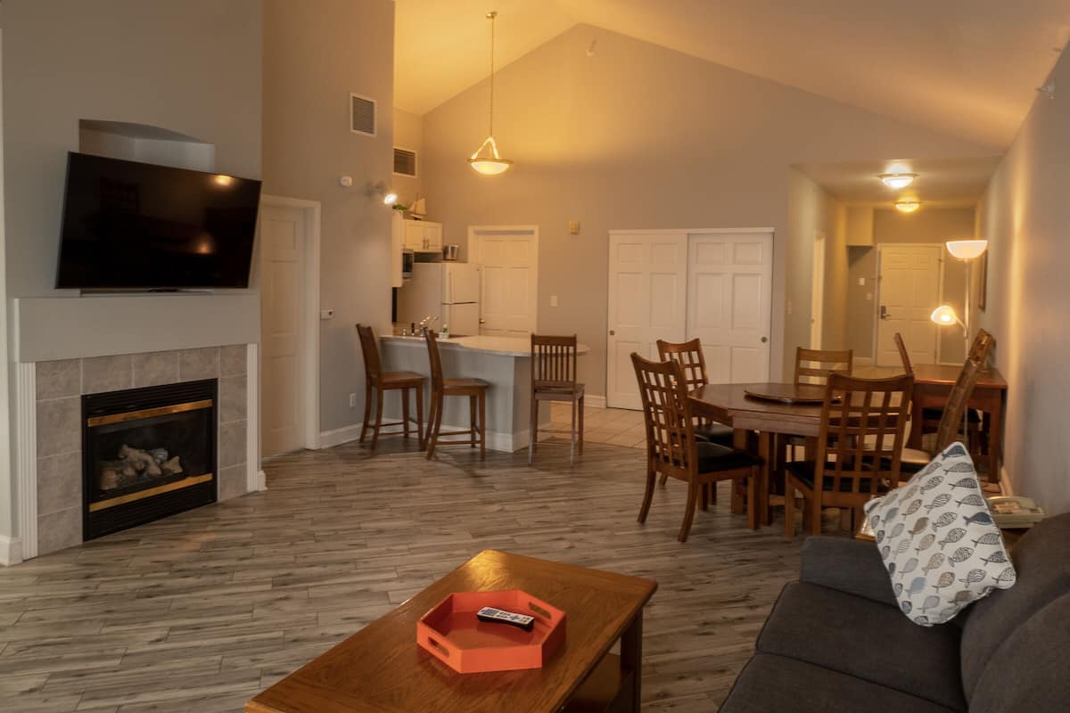 A spacious living room with a fireplace, a dining area with wooden chairs, a kitchen with white cabinets, and a TV mounted on the wall.