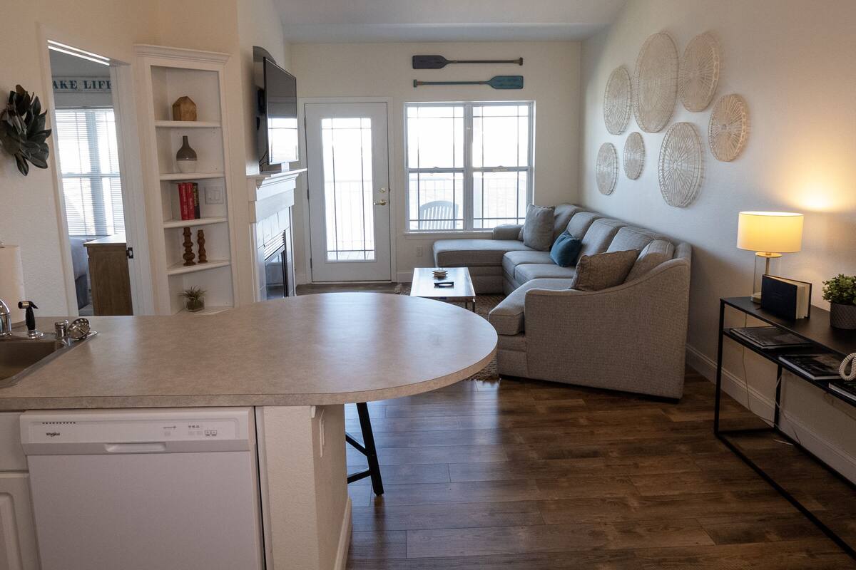 A modern kitchen with a built-in dishwasher, a dining area with a round table, a sofa, and a wall-mounted TV.