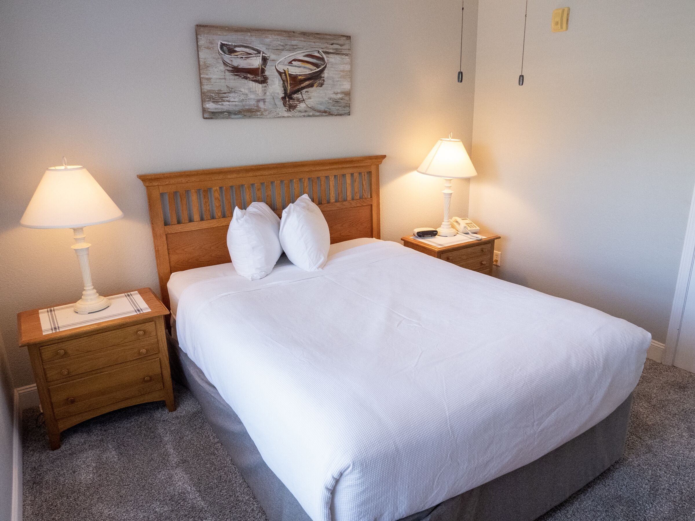 A neatly made bed with white linens and pillows, two bedside tables with lamps, and a painting on the wall.