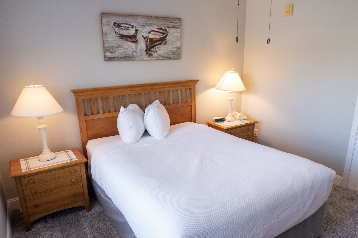 A neatly made bed with white linens and pillows, two bedside tables with lamps, and a painting on the wall.